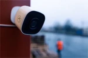 Eine wetterfeste Überwachungskamera mit Weitwinkelobjektiv ist an einer Außenwand montiert und überwacht ein Außengelände bei Regen, im Hintergrund ist eine Person mit Warnweste zu sehen.