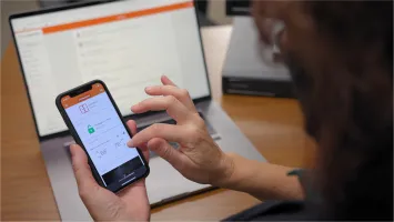 Eine Person bedient die Sicherheits-App von Securitas auf ihrem Smartphone, um Funktionen wie Türverriegelung und Temperaturregelung zu steuern, während ein Laptop im Hintergrund geöffnet ist.