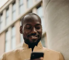 Sicherheitslösungen bequem von unterwegs steuern Ein lächelnder Mann im Mantel steht draußen vor einem modernen Gebäude und sieht zufrieden auf sein Smartphone, bei der Nutzung der Sicherheits-App von Securitas.