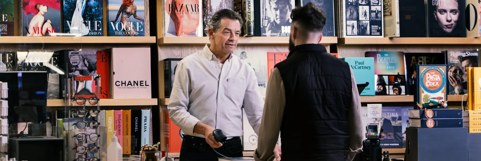 Mitarbeiter im Einzelhandel bedient einen Kunden an der Kasse in einem Buchladen, während Sicherheitslösungen für Unternehmen den Verkaufsraum und den Bezahlvorgang unterstützen.